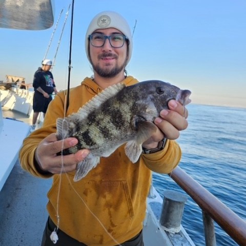 Person on boat holding a fish, wearing a yellow hoodie and beanie, with ocean in the background.