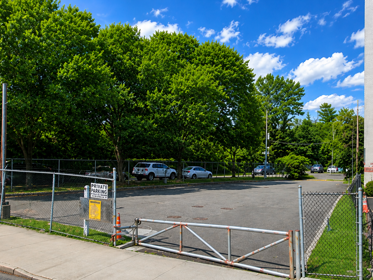 Empty fenced parking lot with trees and cars in the background.