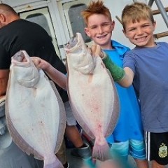 Two smiling boys holding large flat fish on a boat.
