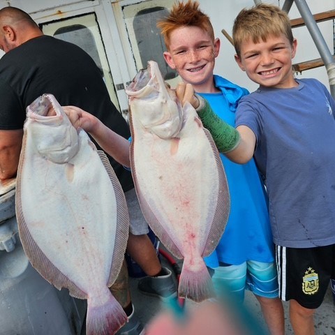 Two boys smiling and holding up two large fish on a boat.