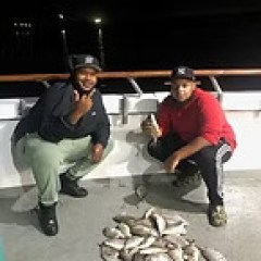Two people on a boat showing off a pile of caught fish at night.