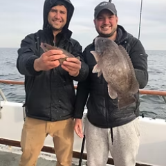 Two men on a boat holding fish, wearing jackets and caps.