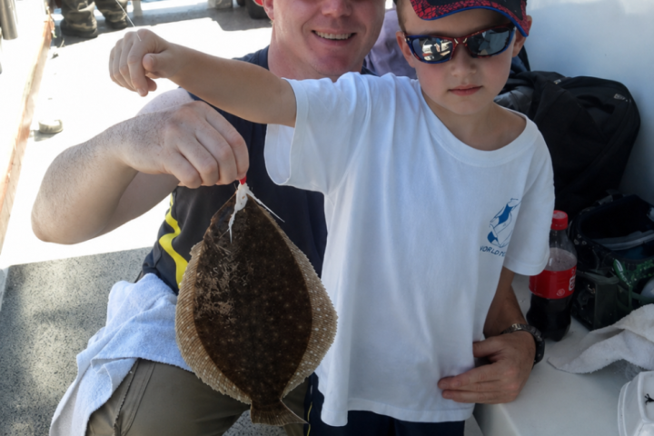 Child holding a flatfish on a boat, assisted by an adult.