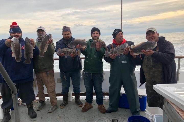 Six people on a boat holding fish with the ocean in the background.